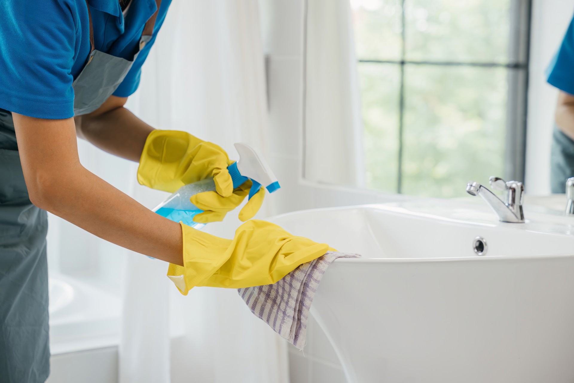 Focused on chores a woman in uniform sprays and cleans bathroom sink and faucet. Her dedication to housework emphasizes purity hygiene and shining fixtures at home. spray cleaner